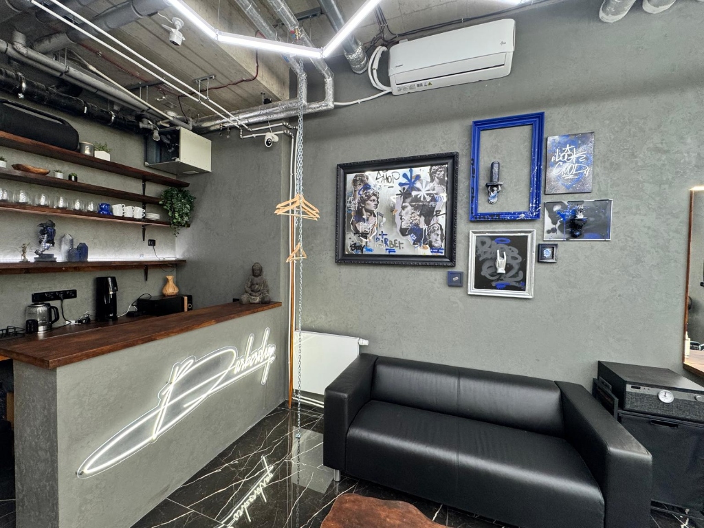 Designová lounge zóna salonu s pohodlnou černou koženou pohovkou, barem a svítícím neonovým nápisem Barbershop